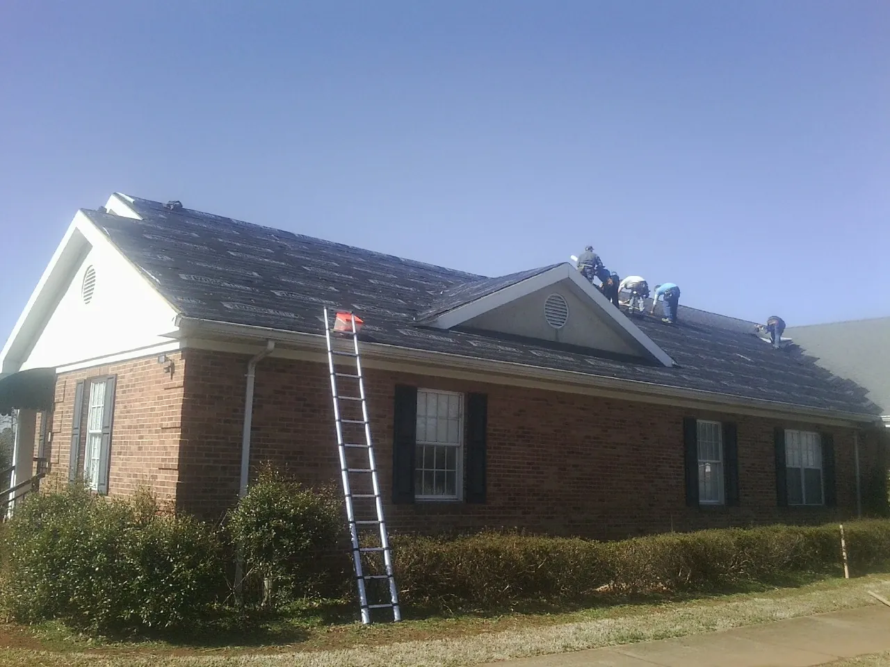 Crew tearing shingles off a brick ranch roof with a ladder set — Metal Roof Repair work by our crew in Wynne