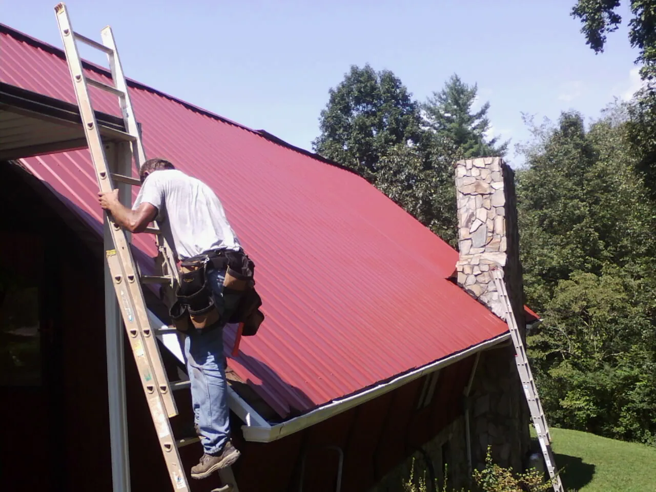 Roofer on a ladder beside a red metal roof and stone chimney — Soffit & Fascia Repair by Roofing Brothers in Wynne